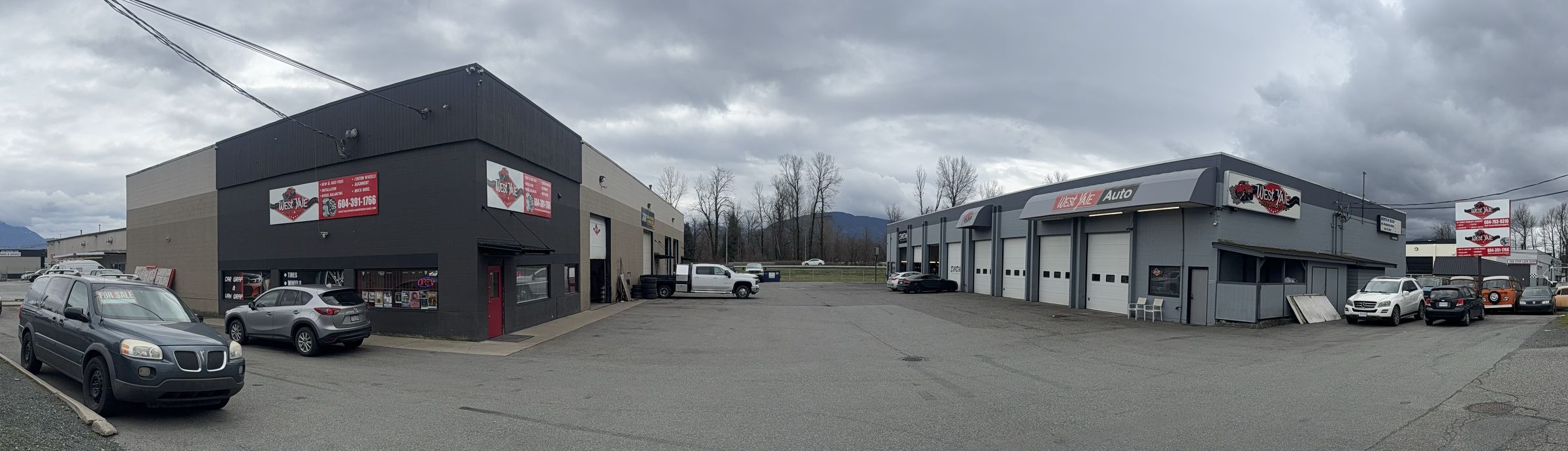 The West Yale lot on Yale Road West: the black Wheels & Tires building on the left, the grey automotive building with bay doors on the right, mountains behind