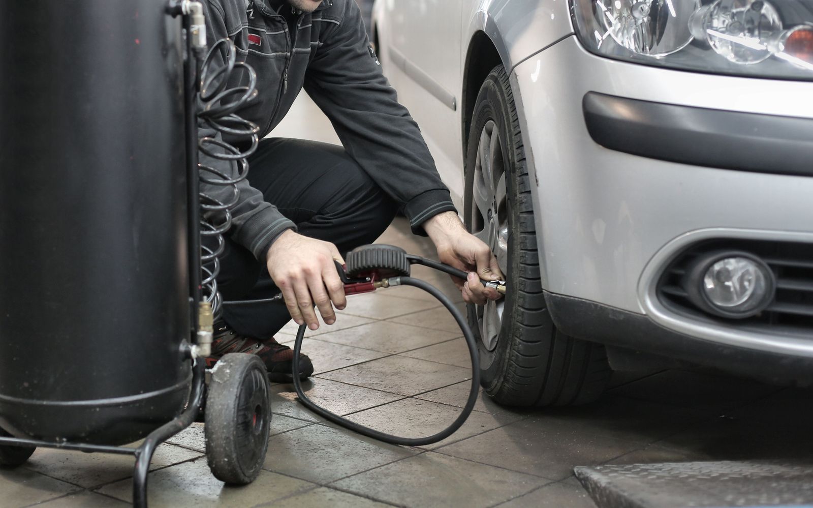 Mechanic crouched beside a silver car's front tire, holding an inflator gauge on the valve stem